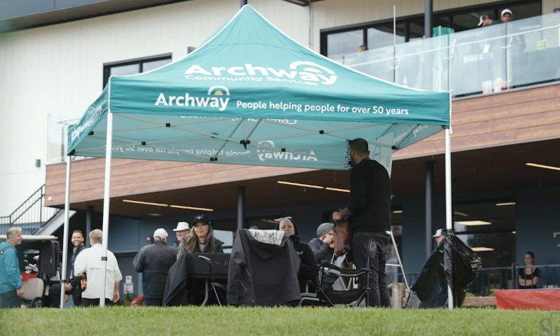Group of people gathered around tables under a teal pop-up canopy, during golf event