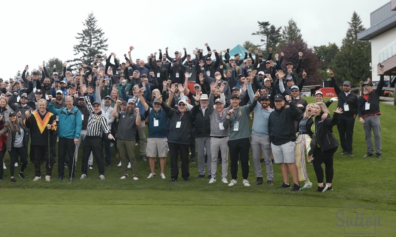 A group of people at a golf charity event stands on the course, raising their hands in celebration